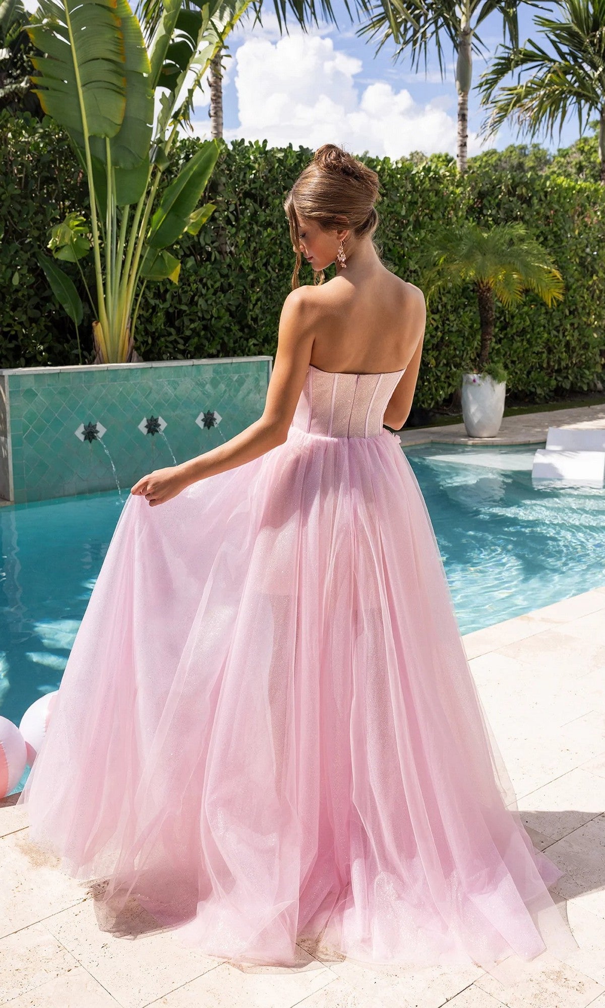 Wearing the Portia and Scarlett PS26078 long formal dress with a glitter tulle skirt, a woman stands by a pool, facing away and holding her gown. Palm trees and lush greenery provide a dreamy backdrop.